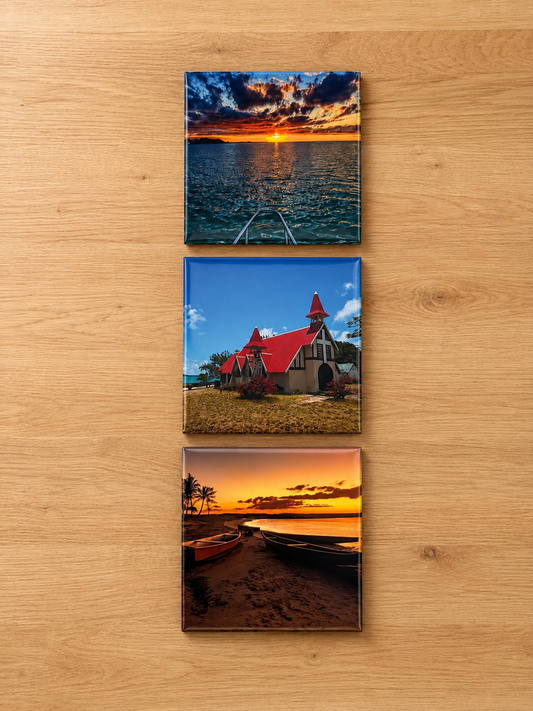 Three vertical square wall art prints showing scenic photographs mounted on a light wood surface. The top print shows a sunset over water with a boat on the horizon, the middle print shows a small church with a red roof, and the bottom print shows a beach scene at sunset with boats on the shore.