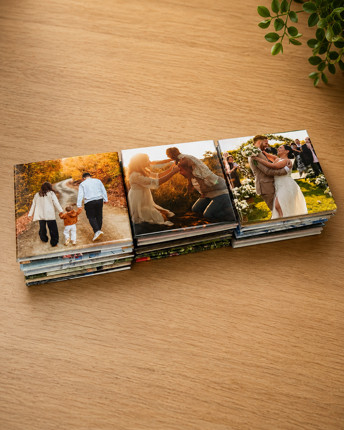A stack of custom square photo prints arranged in a row on a light wood tabletop, with a small green plant leaf visible in the top-right corner. The photos depict family and wedding scenes.