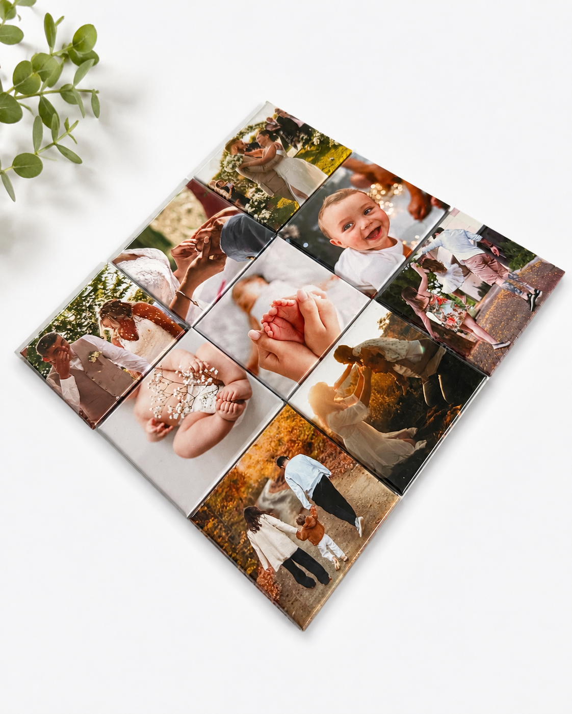 A square custom photo collage print featuring a 3x3 grid of wedding and family photographs, displayed at an angle on a clean white surface with a small green plant leaf in the corner.